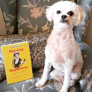 A small white dog sitting on a sofa. Next to her is a book with a yellow cover and the title Hearing Happiness.