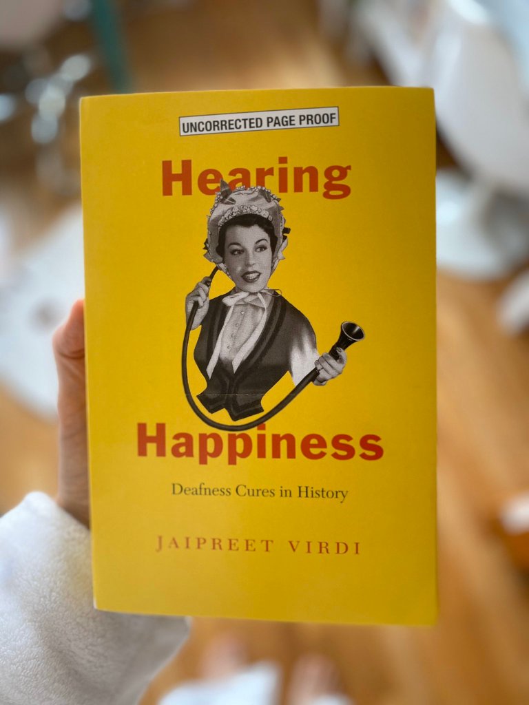 A woman's hand holding the yellow book of Hearing Happiness against a blurry background.