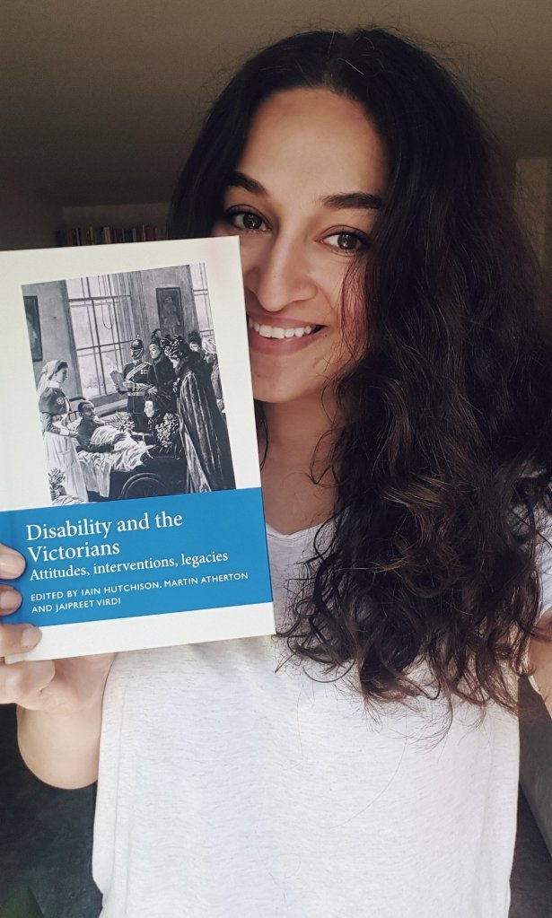 An indian woman with shoulder length curly hair smiling and holding a copy of Disability and the Victorians.