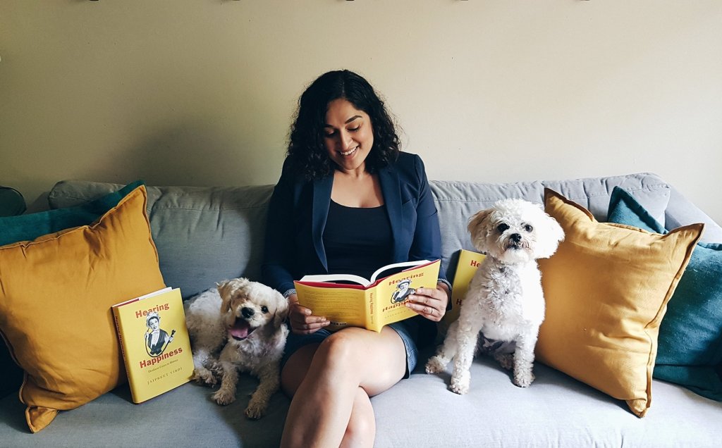 Jai sits in the middle of a grey sofa smiling and looking at her copy of Hearing Happiness. to her right, Benny, a small white dog with one eye, is lying down and leaning against her leg. He is smiling. To Jai's right is Lizzie, a small white dog who is sitting up with a serious expression. There are copies of the book next to both dogs. 